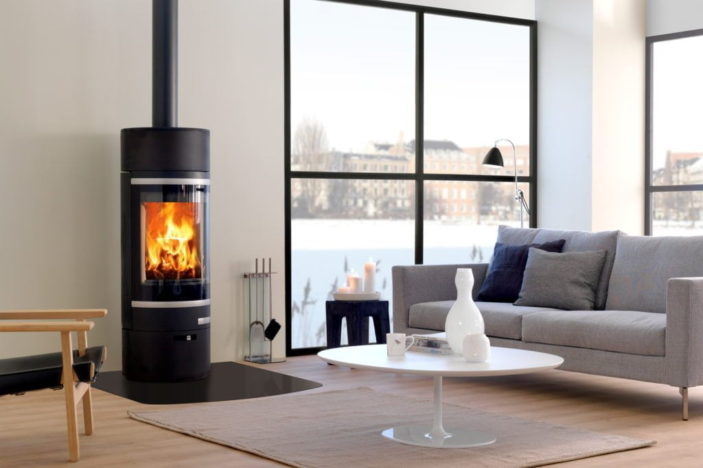 Modern vardagsrum med braskamin, soffa och stora fönster. Eldstadens lågor lyser upp det stilrena rummet, vilket skapar en varm och inbjudande atmosfär.