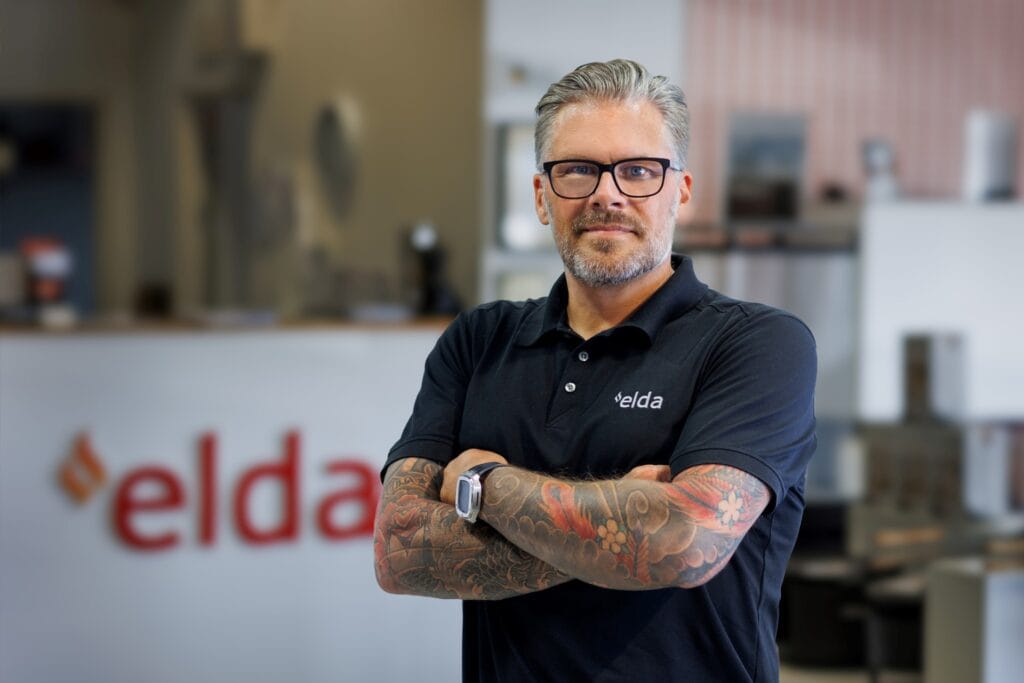 Man i svart pikétröja med "Elda"-logga och tatuerade armar står med armarna i kors framför en suddig bakgrund med "Elda"-skylt.