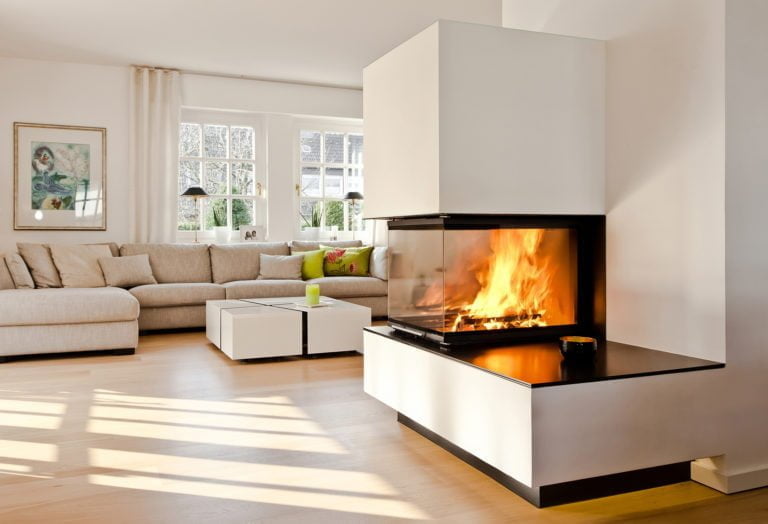 Ljust vardagsrum med modern hörnöppen spis, beige soffa och stora fönster. Elden i spisen skapar en varm och inbjudande atmosfär.