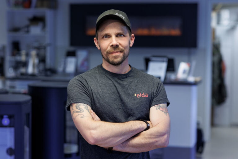 Man i grå t-shirt med "elda" logotyp, står med armarna i kors i en butiksmiljö med en eldstad i bakgrunden.