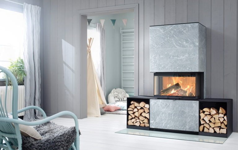 Modern grå stenbeklädd kamin med glasfront och vedförvaring, omgiven av ljus inredning och en blå stol, i ett ljust vardagsrum.