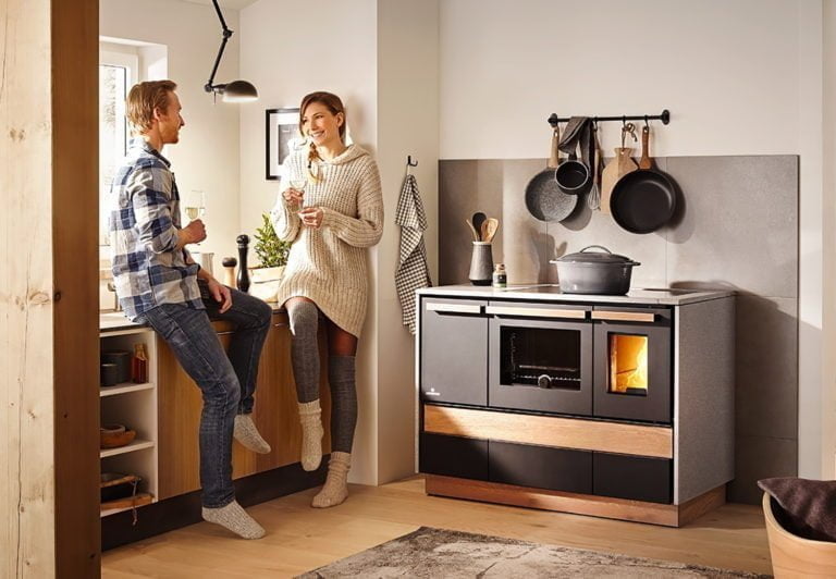 Par i kök dricker vin vid vedspis, omgivna av köksredskap och grytor. Mysig inomhusmiljö med trädetaljer och modern inredning.