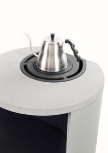 Rund grå kamin med en rostfri stålvattenkokare ovanpå. Kaminen har en svart platta för uppvärmning. Perfekt för utomhusbruk.