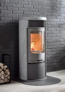 Modern kamin i stål och sten mot en grå tegelvägg, med brinnande eld inuti. Vedförvaring syns bredvid kaminen.