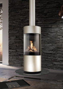 Modern rund eldstad med glasfront i ett rum med stenväggar. Elden brinner inuti, och en surfplatta ligger på golvet.