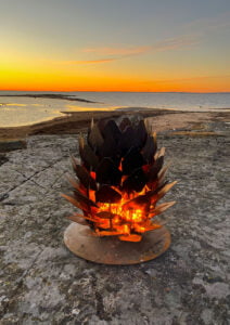 Eldkorg i metall brinner på en klippa vid havet under en solnedgång, skapar en varm atmosfär med orange himmel och vatten i bakgrunden.