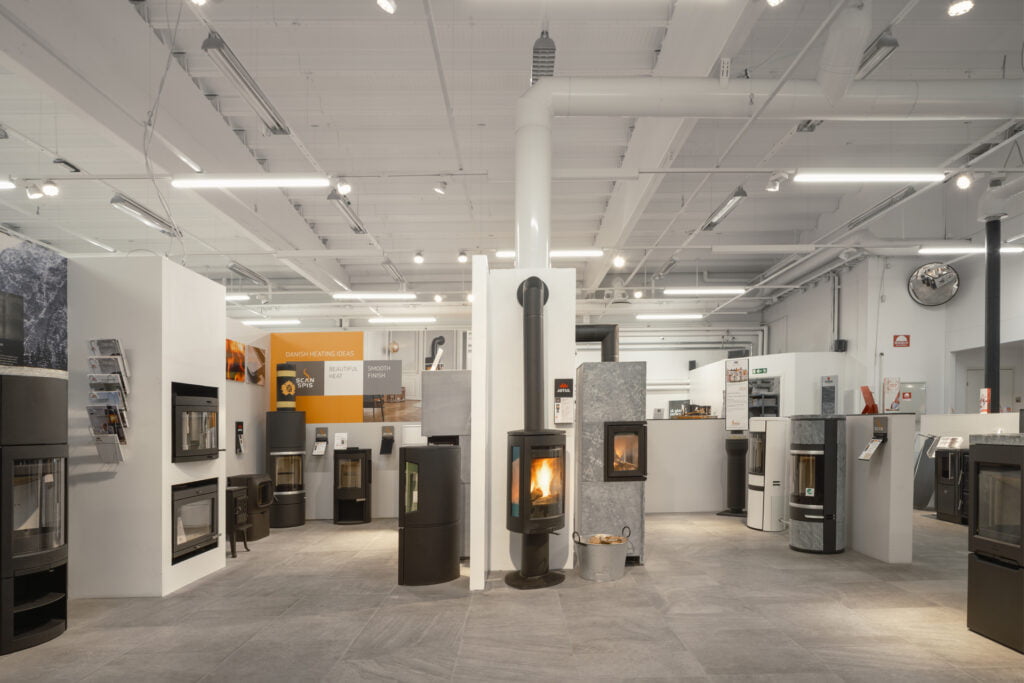Utställningshall med moderna eldstäder och kaminer, upplyst av taklampor. Eldstäderna är placerade på rad i ett välorganiserat showroom.