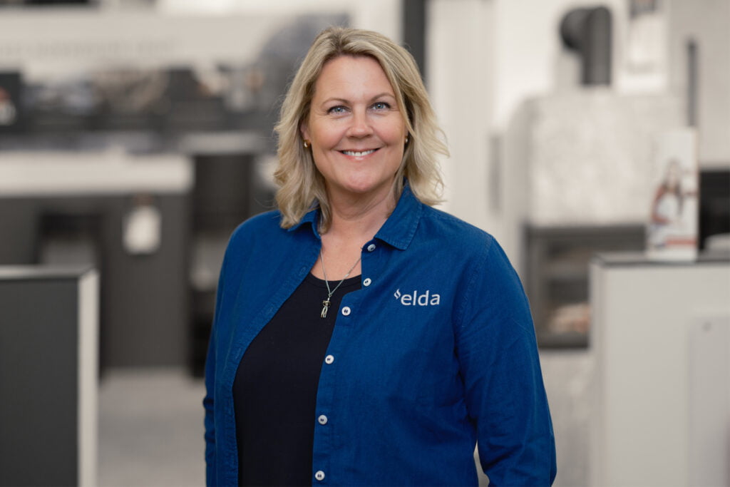 Kvinna i blå "Elda"-skjorta står i en butiksmiljö, leende mot kameran. Bakgrunden visar suddig inredning och skyltar.