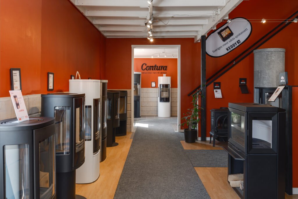 Utställning av moderna kaminer i butik med orange väggar, inklusive märkena Contura och Jotul. Ljus trä- och grå matta på golvet.