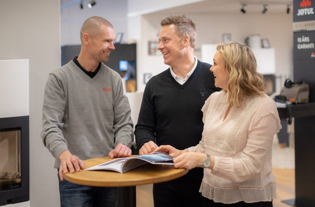 Tre personer diskuterar vid ett runt bord inomhus. En broschyr ligger på bordet. Bakgrunden visar en ljus, modern butiksmiljö med eldstad och Jotul-skylt.