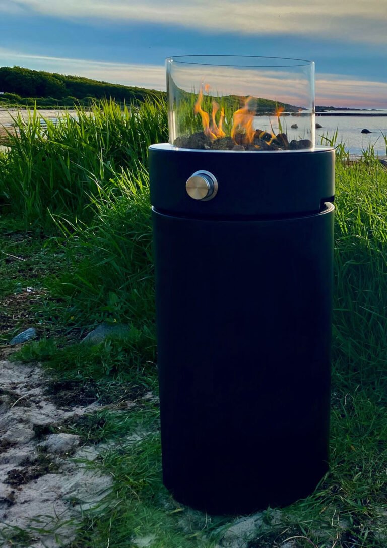Svart eldstad med glasbehållare och synliga lågor, placerad på en grönskande strand vid havet under en blå himmel.