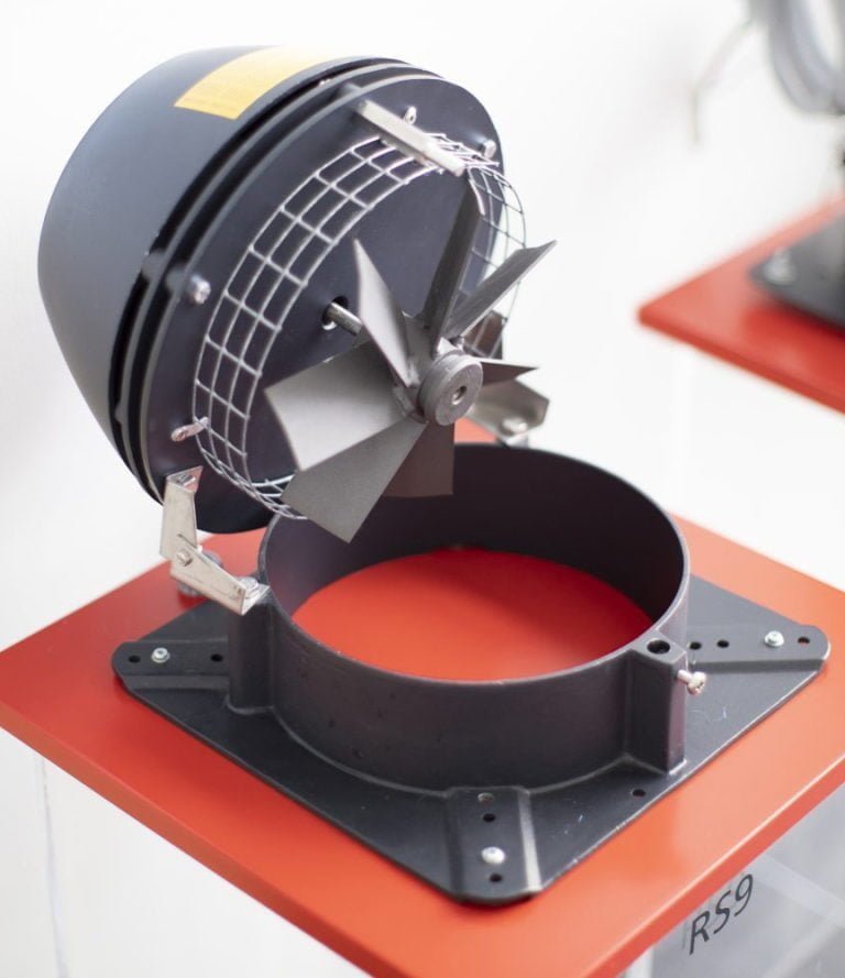 Svart och röd ventilationsfläkt monterad på en röd display. Fläkten har metallblad och ett skyddsgaller, placerad på en röd yta.