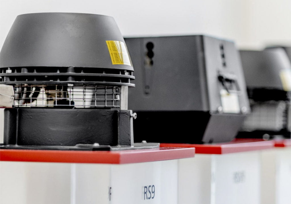 Svart ventilationssystem på rad, monterat ovanpå vita enheter med röda toppar. Fokus på närmaste ventilationsenhet.