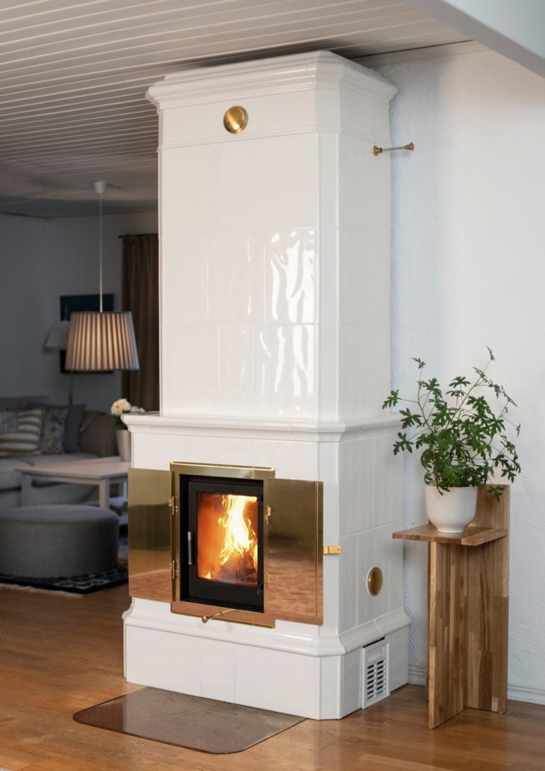 Vit kakelugn med brinnande eld, guldfärgade detaljer och en krukväxt på ett träbord bredvid. Mysig inomhusmiljö med soffa och lampa i bakgrunden.