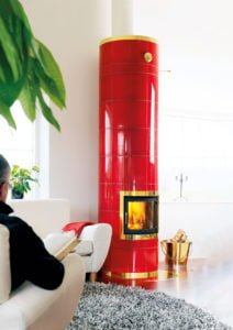 Person sitter i en vit fåtölj bredvid en röd, kaklad kamin med eld. Guldfärgad vedhink och grå matta i förgrunden.