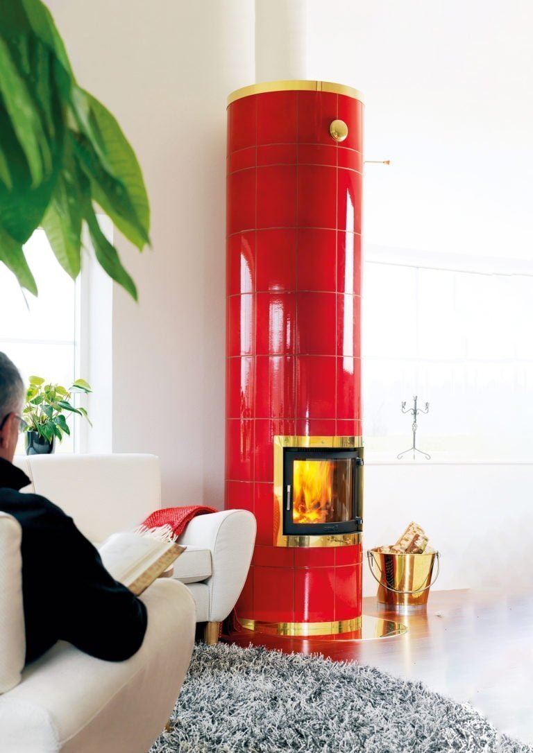 Person sitter i en vit fåtölj bredvid en röd, kaklad kamin med eld. Guldfärgad vedhink och grå matta i förgrunden.