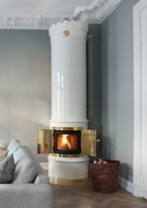Vit kakelugn med guldfärgad eldstadsdörr, brinnande ved inuti. Grå väggar och soffa i ett elegant vardagsrum. Brunt vedförvaringskärl på golvet.
