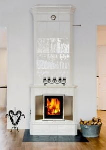 Vit kakelugn med brinnande eld, dekorerad med ljusstakar. Vedförvaring i en metallhink bredvid. Trägolv i ett ljust rum.