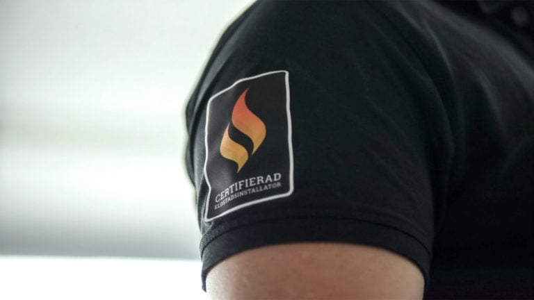 Svart tröja med märke för certifierad eldstadsinstallatör, visar en flamlogotyp i orange och guldfärg.