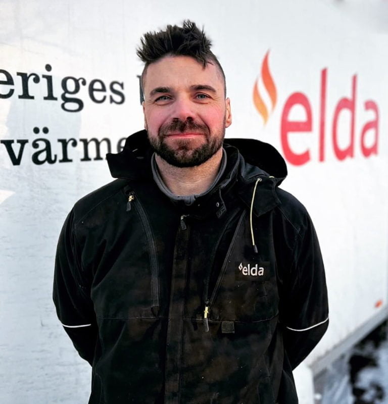 Man i svart jacka står framför vit vägg med "elda"-logotyp. Snöigt underlag syns i bakgrunden.