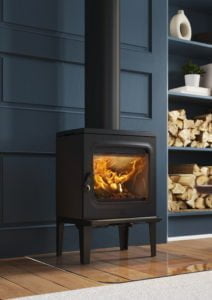 Svart vedeldad kamin med brinnande eld, placerad mot en blå panelvägg. Vedstapel och prydnadssaker syns på en hylla bredvid.