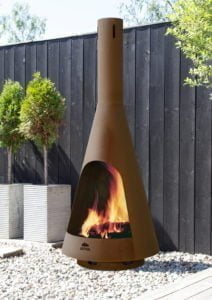 Uteplats med en modern utekamin från Jøtul, omgiven av grus och träd, mot en mörk trävägg. Elden brinner i kaminen.