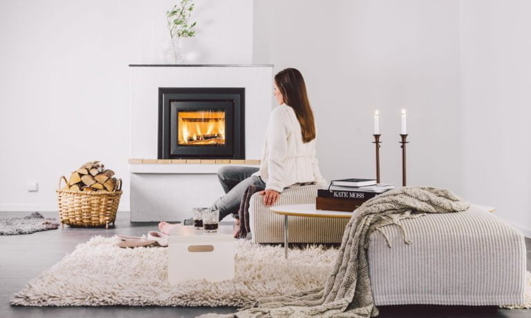 Kvinna sitter framför en modern eldstad i ett stilrent vardagsrum. Ved i korg, tända ljus och böcker skapar en mysig atmosfär.