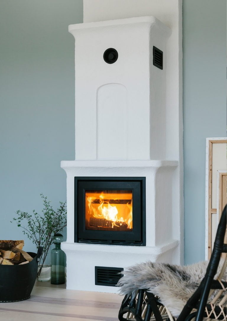 Vit eldstad med brinnande ved, omgiven av en vedkorg och en fåtölj med fårskinn. Ljusgrön vägg i bakgrunden.