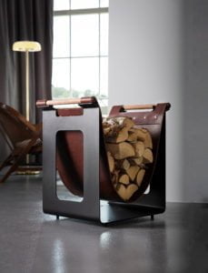 Elegant vedkorg i metall och läder fylld med ved, placerad på ett modernt golv framför ett stort fönster med gardin och lampa.