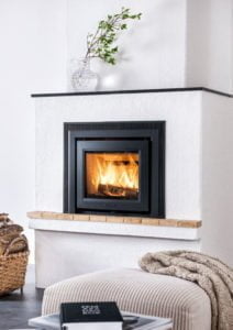 Modern braskamin med brinnande eld, omgiven av vit putsad vägg. En korg med ved och en dekorativ kvist i glasvas finns i närheten.