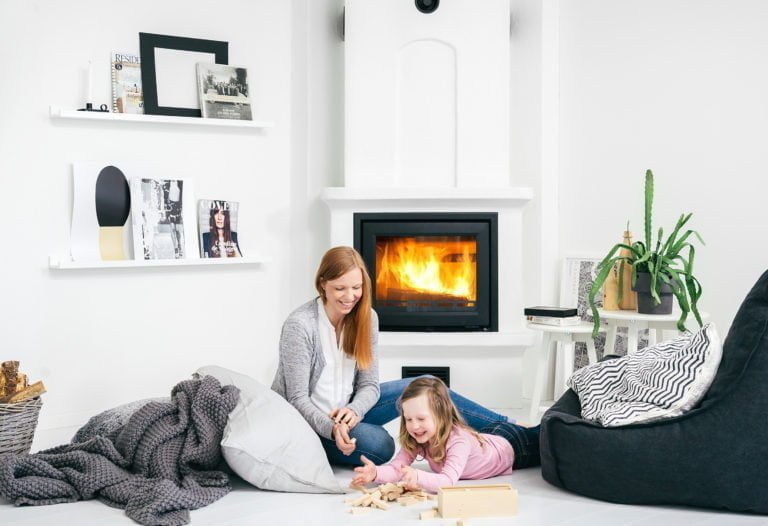 Kvinna och barn leker vid en brinnande kamin i ett modernt vardagsrum. Hyllor med böcker och dekorationer syns i bakgrunden.