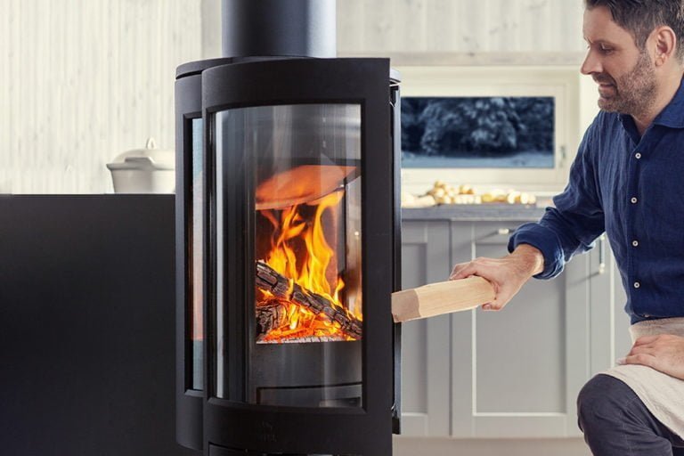 Man lägger ved i en modern kamin med glaslucka, omgiven av ett ljust kök. Elden brinner klart och varmt. Nyckelord: kamin, eld.