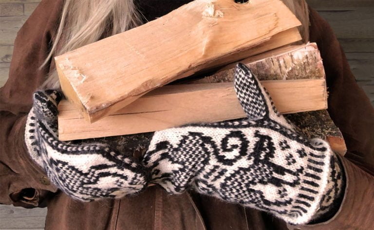 Person klädd i brun jacka och stickade vantar håller vedträn.