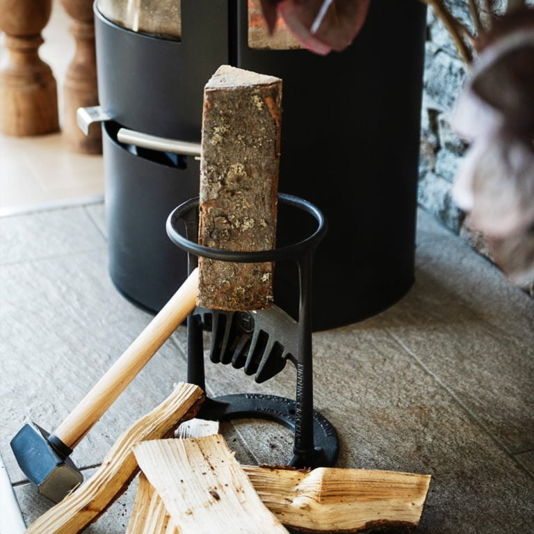 Vedklyvningsverktyg och yxa bredvid vedträn på golv, intill en svart vedkamin.