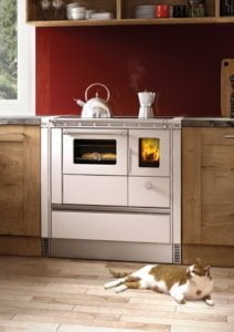 Vedeldad spis med brinnande eld, bröd i ugnen och tekanna ovanpå, placerad i ett kök med röd vägg. Katt ligger på golvet framför spisen.