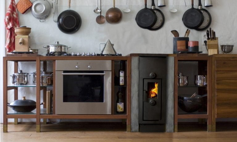 Modernt kök med vedspis, ugn och köksredskap på väggen. Trähyllor med kastruller och flaskor. Brinnande eld i spisen ger en mysig atmosfär.