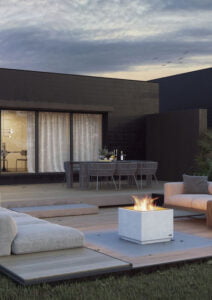Modern uteplats med eldstad, stilrena möbler och matplats framför ett mörkt hus. Mysig kvällsstämning under molnig himmel. Perfekt för avkoppling utomhus.