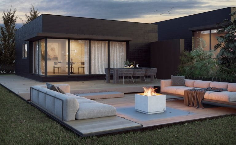 Modern uteplats med eldstad, soffor och matbord. Glasfönster visar inomhusbelysning och dekoration. Omgiven av grönska och kvällshimmel. Perfekt för avkoppling utomhus.