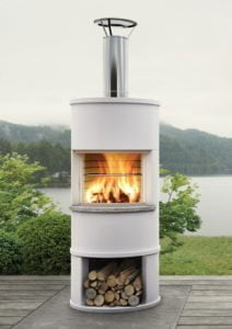 Utomhuskamin med brinnande eld och vedförvaring, placerad på en terrass med sjö och skog i bakgrunden. Perfekt för trädgården eller uteplatsen.