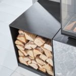 Vedförvaring i svart metallställ bredvid en kamin. Staplade vedträn i en modern, stilren design på ljust golv.