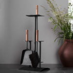 Svart eldstadsset med borste och skyffel med trähandtag på grå bakgrund, bredvid en brun vas med gröna växter.