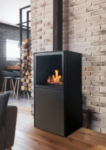 Modern svart eldstad mot vit tegelvägg, med eld inuti. Träbitar staplade bredvid och stilren inredning i bakgrunden. Perfekt för hemtrevlig atmosfär.