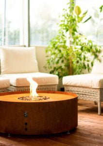 Rund eldstad i rostbrunt material på träaltan, omgiven av vita korgmöbler och grön växt. Elden brinner lugnt i mitten.