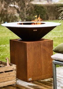 Rostfärgad eldstad på trätrall, omgiven av grönska. Elden brinner i en skålformad behållare ovanpå en rektangulär bas.