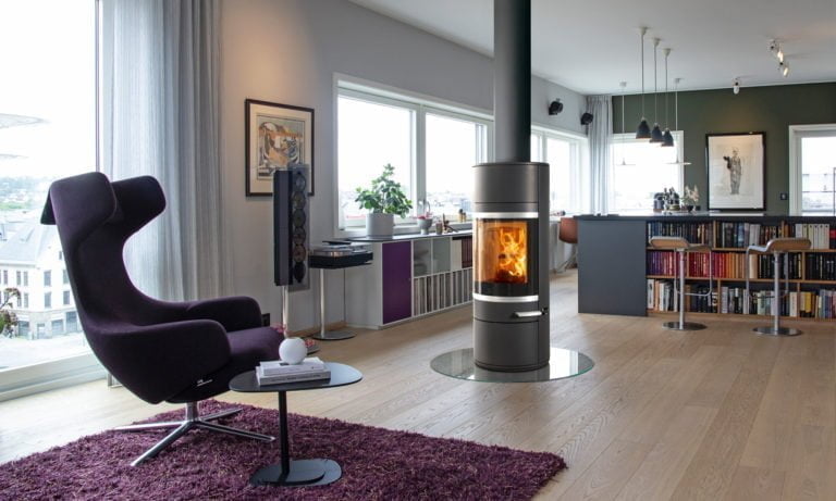 Modernt vardagsrum med en braskamin i centrum, omgiven av en fåtölj, bokhylla och köksbar. Stora fönster släpper in naturligt ljus.