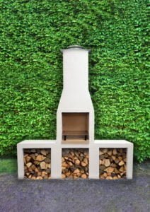 Vit utomhusgrill med skorsten, omgiven av grön häck. Vedförvaring under grillen. Perfekt för trädgårdsgrillning och utomhusliv.