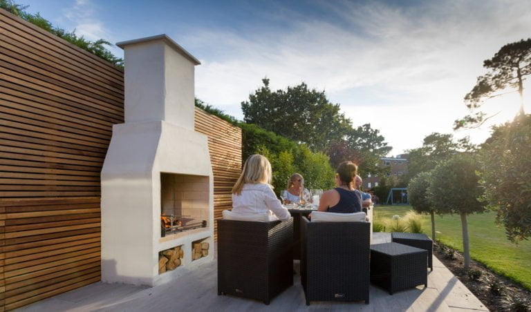Alt-text: Utomhusmiljö med personer som sitter vid ett bord nära en vit utomhusspis, omgivna av träpanel och grönska. Solnedgång i bakgrunden.