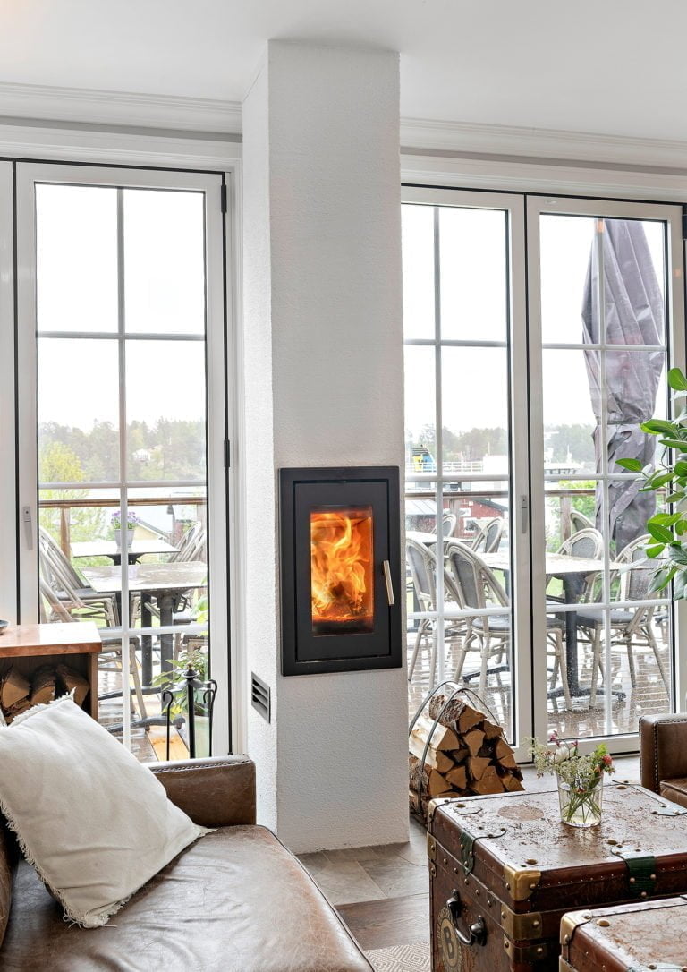 Modernt vardagsrum med braskamin, stora fönster och utsikt mot en möblerad altan. Mysig interiör med lädersoffa och växt. Nyckelord: braskamin, vardagsrum.