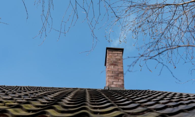 Röda tegelstenar bildar en skorsten på ett tak med svarta takpannor. Blå himmel och nakna trädgrenar syns i bakgrunden.