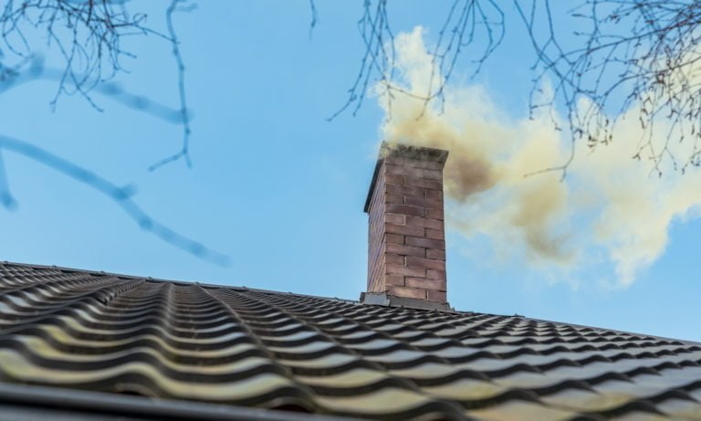 Tegelstensskorsten på hustak släpper ut rök mot en klarblå himmel, omgiven av kala trädgrenar. Skorsten och rök är i fokus.